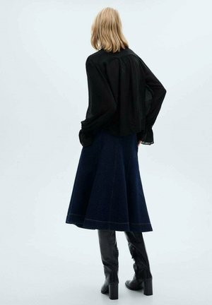 Blusa nera trasparente a maniche lunghe abbinata a una gonna lunghezza ginocchio in denim scuro svasato. Stivali neri alti fino al ginocchio completano l'outfit, evidenziando il contrasto di texture.