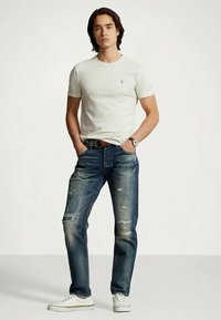 Bílé tričko s krátkým rukávem, vybledlé modré džíny s ošoupanými místy, opotřebovaná textura a bílé tenisky. Model stojí s rukama v kapsách.