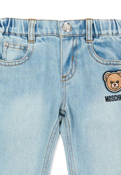 Pantalones de mezclilla azul claro para niños con cintura elástica, bolsillos delanteros, botón de metal y un parche de osito con el texto "Moschino" en el lado derecho.