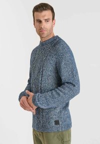 Blå stickad tröja med ett texturerat mönster och hög krage. Har ribbade ärmslut och nederkant, tillverkad av en blandad garnblandning.