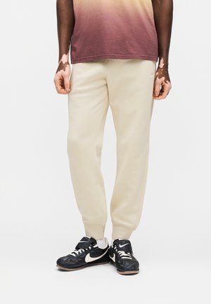 Osoba ubrana w beżowe spodnie dresowe, czarno-białe buty Nike oraz wyblakły brązowo-beżowy gradientowy t-shirt, stoi na tle białego tła.