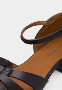 Μαύρο δερμάτινο σανδάλι με λεπτές λουράκια και χαμηλό τακούνι, με ταμπά εσωτερική σόλα και λουράκι στον αστράγαλο με κούμπωμα.