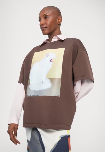 Brązowy, oversize, sweatshirt z grafiką kota na przodzie. Nałożony na jasnoróżową koszulę z kołnierzykiem. Gładka tekstura materiału.