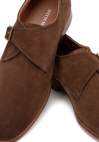 Zapatos de ante marrón con puntera redondeada y una solapa en la parte delantera, asegurada con una hebilla dorada; presenta costuras visibles y un tacón de madera.