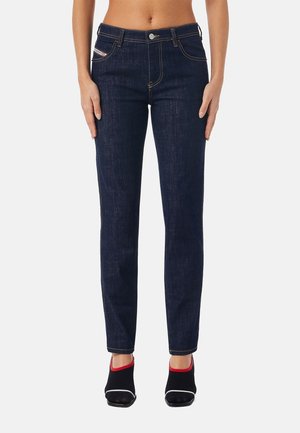 Dunkelblaue Slim-Fit-Jeans, getragen von einer Person mit freiem Bauch und schwarzen Slip-On-Schuhen mit rotem Besatz und weißen Sohlen.