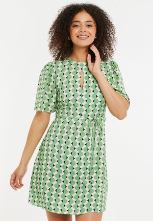 Vihreä ja valkoinen kuviollinen mekko, jossa on lyhyt puffihihnat, avainaukko kaula-aukko ja solmittava vyötärö. Mekossa on geometrinen muotoilu, jossa on päällekkäisiä ympyröitä.