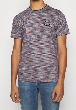 Homme portant un T-shirt à manches courtes rayé horizontalement multicolore avec un petit visage souriant noir sur la poitrine gauche, assorti à un jean bleu.