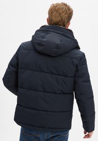 Marine gewatteerde jas met capuchon, voorzien van een textuurafwerking en horizontale stiksels. Achteraan gezien toont hij een relaxte pasvorm en geen zichtbare branding.