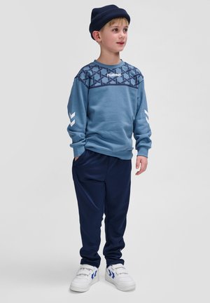 Blauwe sweatshirt met marineblauwe schouderpatroon, wit logo en witte chevrons. Marineblauwe broek en witte schoenen met blauwe accenten maken de outfit compleet.
