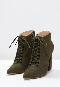 Botines de ante verde oliva con punta afilada, diseño de cordones en la parte delantera y un tacón alto cuadrado. Cierre lateral con cremallera para facilitar su uso.