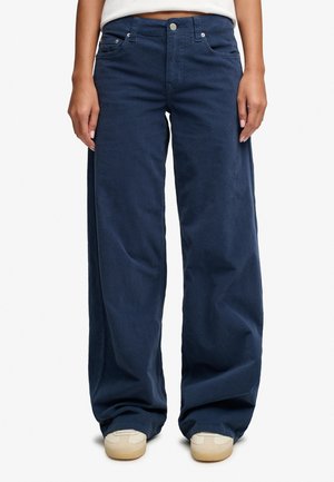 Persona con pantalones anchos azul marino y zapatillas beige de pie sobre un fondo blanco.
