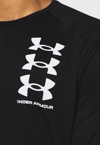 Czarna koszulka z długim rękawem z trzema białymi logo Under Armour i tekstem na środku, wykonana z gładkiego syntetycznego materiału.