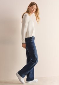 Witte longsleeve top met een gladde textuur, gecombineerd met donkerblauwe jeans en witte sneakers, van de zijkant bekeken tegen een neutrale achtergrond.