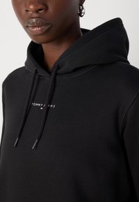 Sudadera con capucha negra, hecha de una mezcla de algodón, con un pequeño logo blanco "TOMMY JEANS" en el pecho y cordones con puntas de metal.