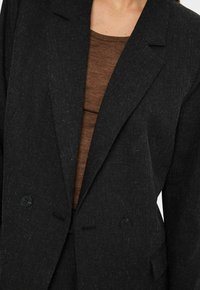 Primo piano di una persona che indossa un blazer doppiopetto grigio scuro sopra un top a girocollo marrone.