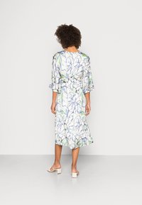 Vestido midi floral em branco, com padrões verdes e azuis, mangas soltas e uma cintura ajustada para maior forma. Sandálias de salto quadrado.