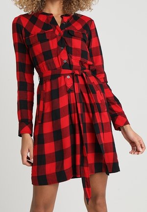 Femme portant une robe-chemise à carreaux rouge et noir, manches longues, boutons à l'avant, poches poitrine et ceinture nouée à la taille.