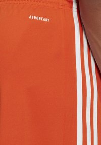 Orangene Sportshorts aus leichtem Material, mit drei weißen vertikalen Streifen und einem "AEROREADY"-Logo am Bund.