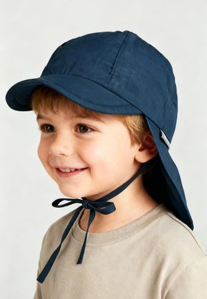 Smiling young boy wearing a navy sun hat with neck flap and chin ties, dressed in a beige shirt, against a plain background.