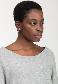 Maglione grigio a maglia con scollatura ampia, caratterizzato da una texture morbida e delicati luccichii. Il modello ha i capelli corti e indossa piccoli orecchini.