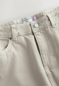 Jupe en denim beige avec passants pour ceinture, bouton en métal gravé de "NEXT DENIM SINCE 1982" et une étiquette visible portant l'inscription "HOURGLASS SKIRT".