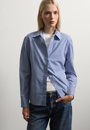 Chemise à carreaux bleus et blancs avec col, manches longues et coupe décontractée, superposée sur un haut blanc ajusté.