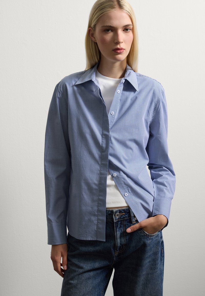Camisa de cuadros en azul y blanco con cuello, mangas largas y un ajuste relajado, superpuesta sobre una blusa blanca ajustada.