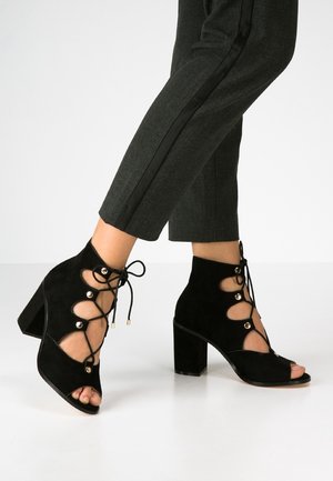 Czarne zamszowe botki na wysokim obcasie, sięgające do kostki, z otwartym noskiem, posiadają sznurowane z przodu z złotymi akcentami oraz wycięciami.