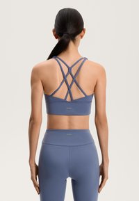 Športová podprsenka v tlmenej modrej farbe s prekríženými ramienkami na chrbte. Hladká textúra s jemným brandingom na dolnom okraji.