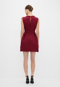 Abito senza maniche di colore bordeaux con corpetto fitted, gonna ampia e chiusura a chiave sulla schiena. Abbinato a scarpe nere con tacco alto.