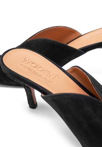 Svarta mocka klackmules med öppen bakdel, med en beige innersula präglad med "WODEN." Klacken har en texturerad detalj för bättre grepp.