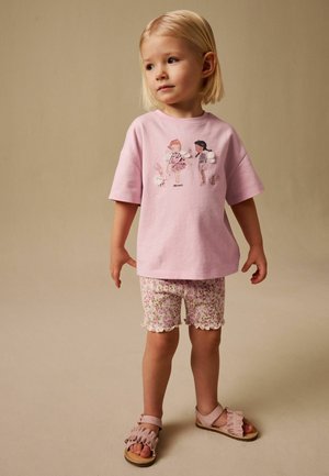 Junges Kind mit blondem Haar, das ein rosa T-Shirt mit Feenillustrationen, geblümte Shorts und rosa Sandalen trägt, auf einem beigen Hintergrund stehend.