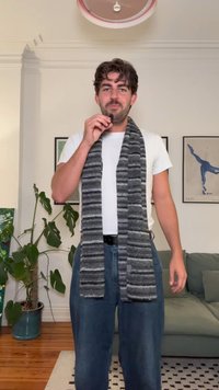 Écharpe rayée grise et noire en tissu tricoté, drapée autour du cou. Portée avec un t-shirt blanc et un jean bleu, debout à l'intérieur.