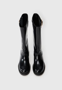 Botas negras hasta la rodilla hechas de material brillante con puntera redondeada y un sutil detalle de correa. Textura suave y suela resistente.