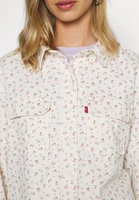 Chemise imprimée florale avec un fond crème, présentant des fleurs roses et vertes, deux poches avant et une étiquette logo Levi's rouge.