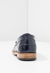 Mocassin en cuir bleu marine brillant avec coutures visibles, talon empilé en bois et semelle noire, vu de dos sur une surface blanche.