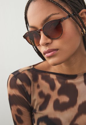 Femme aux cheveux tressés portant des lunettes de soleil écaille de tortue et un haut transparent à imprimé léopard, regardant légèrement vers le bas sur un fond uni.