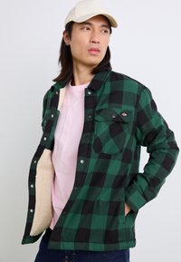 Veste à carreaux verts et noirs avec une doublure douce crème, dotée de boutons-pression et de poches poitrine, portée par-dessus une chemise rose.