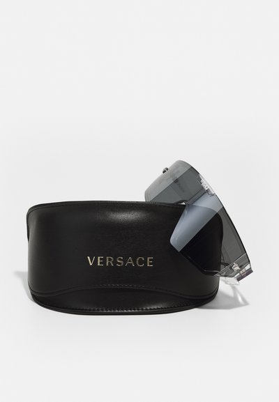 Black leather eyewear case with gold "VERSACE" logo, holding oversized gray sunglasses with clear accents and geometric lenses.