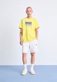 Yellow cotton t-shirt with graphic print, paired with white shorts featuring an Adidas logo. Worn with light sneakers and white socks.