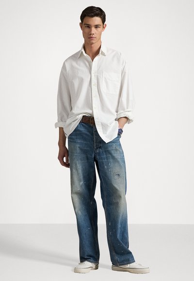 Camisa blanca de botón con mangas remangadas, jeans de mezclilla con un lavado azul desgastado y detalles desgastados, combinados con zapatos blancos.