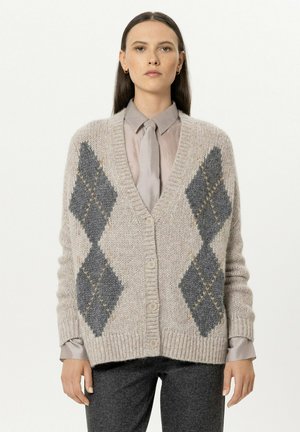 Woman wearing a beige knitted cardigan with large gray diamond patterns, a taupe shirt, and a matching tie, standing against a white background.