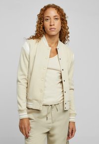 Beige bomberjakke med knappeåpning, ribbede mansjetter og side-striper. Brukt over en matchende lys topp. Teksturert stoff og avslappet passform.