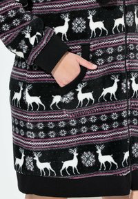 Giacca in pile nera con un design a motivo che presenta renne bianche e fiocchi di neve; include una chiusura con zip e tasche laterali con polsini neri.