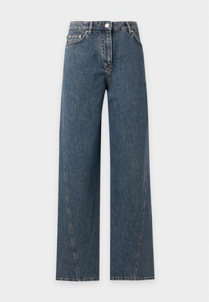 Mujer con cabello largo y rizado, vestida con un suéter de cuello alto gris oscuro y jeans azules de talle alto y holgados, de pie contra un fondo claro y liso.
