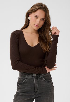 Femme aux longs cheveux bruns porte un haut à manches longues marron foncé à col en V, un jean foncé et de grandes boucles d’oreilles en argent, debout devant un fond uni.
