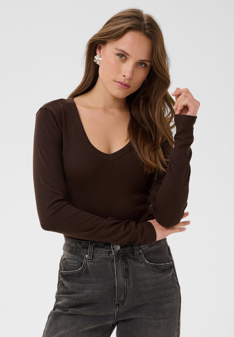 Femme aux longs cheveux bruns porte un haut à manches longues marron foncé à col en V, un jean foncé et de grandes boucles d’oreilles en argent, debout devant un fond uni.