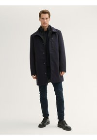 Abrigo de lana azul marino con cierre de botón, gran cuello y bolsillo en el pecho con cremallera, combinado con pantalones azul marino y zapatos negros de cordones.