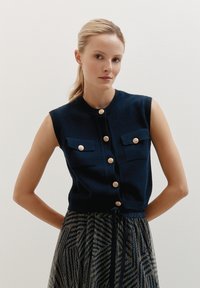 Marineblaues, ärmelloses Strickoberteil mit zwei Fronttaschen, goldenen Knöpfen und tailliertem Design, kombiniert mit einem gemusterten Rock.