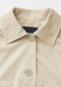 Colletto e parte superiore anteriore di un cappotto beige con due bottoni, cuciture visibili e una targhetta nera parzialmente visibile all'interno del collo.